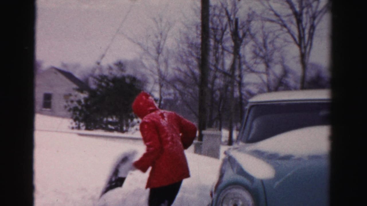 Person in a red coat tossing snow from a shovel near the front of a parked car.