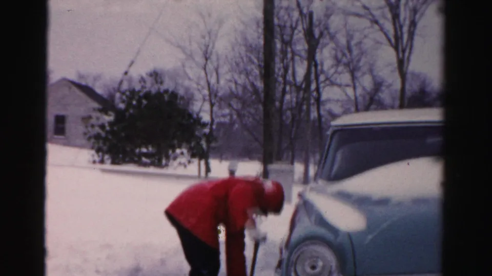 The bundled person bends low with the shovel while clearing snow near a vintage car in Albany winter footage.