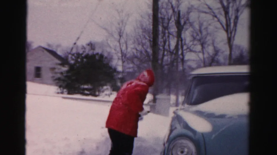 A red-coated figure continues shoveling snow near a parked car in a quiet Albany, New York neighborhood.