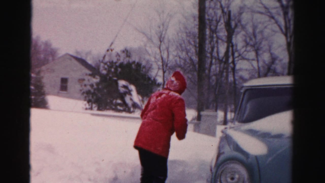 In Albany winter home-movie footage, a person in a bright red coat shovels deep snow beside a vintage car in a residential yard.