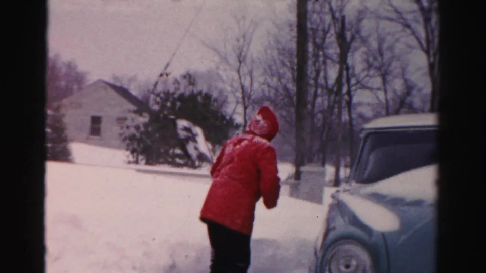 In Albany winter home-movie footage, a person in a bright red coat shovels deep snow beside a vintage car in a residential yard.