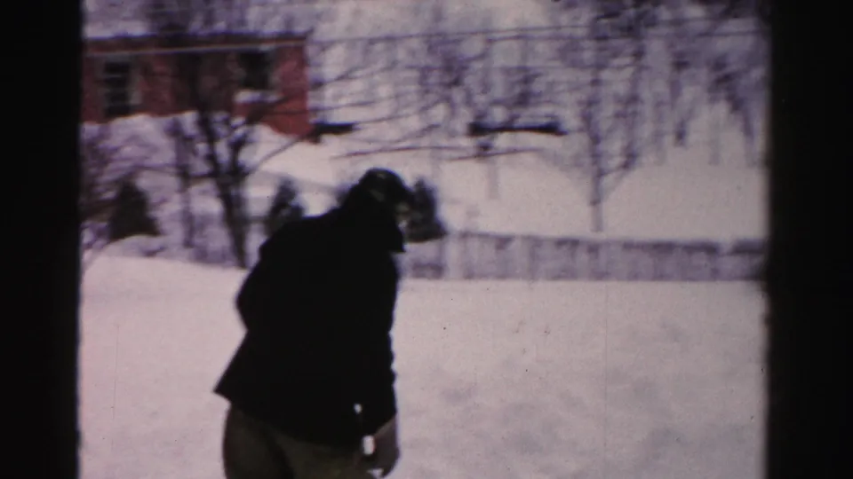 A bundled person leans into the deep snow in an Albany yard, with a house, bare branches, and snowbanks visible behind them.