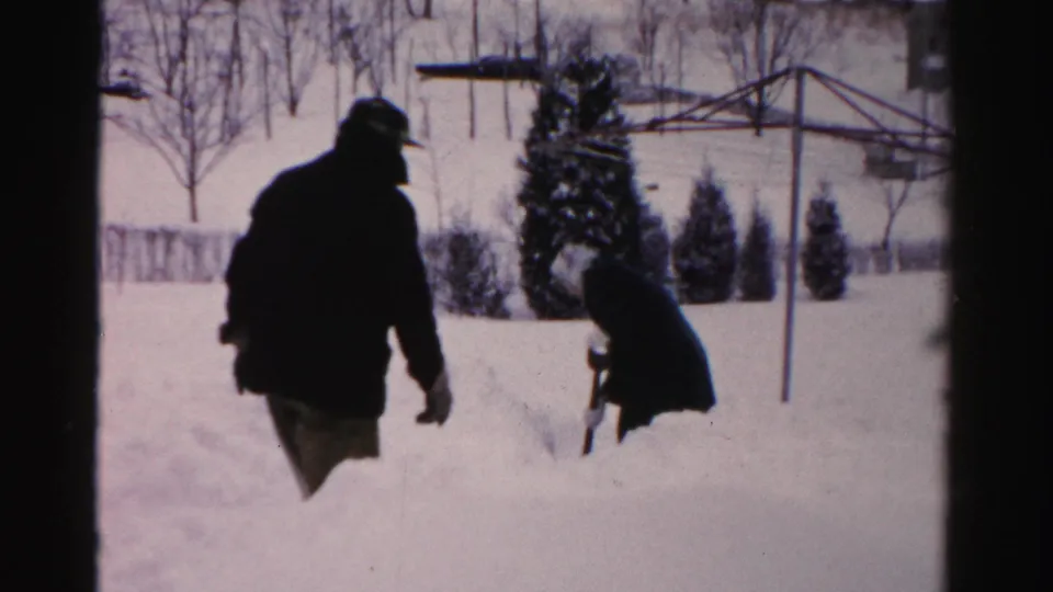 Two bundled figures appear to clear or move through deep snow in an Albany yard, with a clothesline, shrubs, and houses in view.