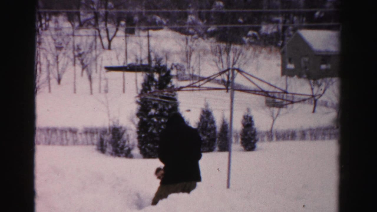 In a 1950s Albany backyard, an adult in a dark winter coat moves through deep snow near evergreen shrubs and a metal clothesline.