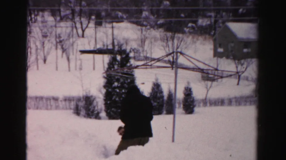 In a 1950s Albany backyard, an adult in a dark winter coat moves through deep snow near evergreen shrubs and a metal clothesline.