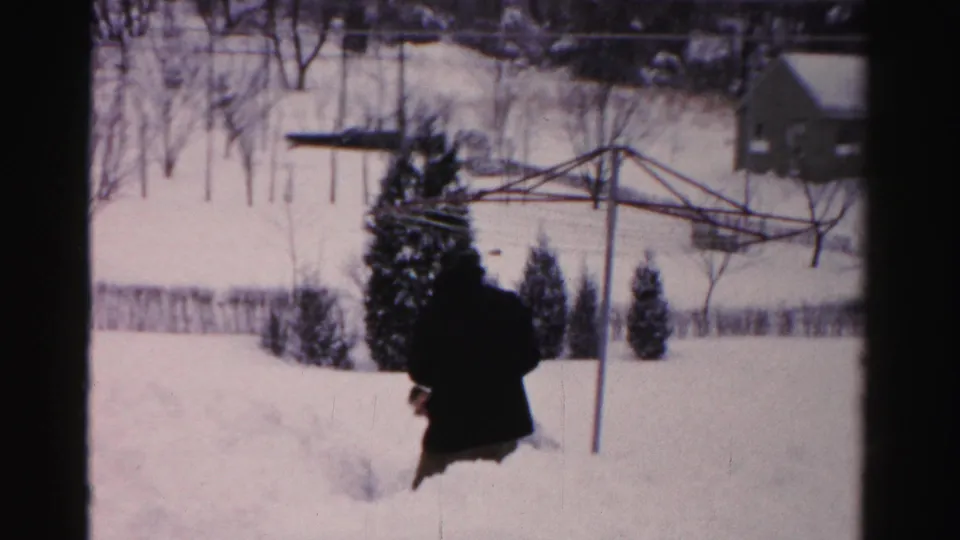 An adult in dark winter clothing walks through deep snow in an Albany residential yard, with a clothesline frame and small outbuilding visible.