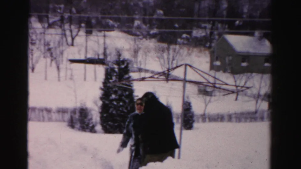 Two adults appear close together in the snowy Albany yard, one in a dark coat and another partly visible near the evergreen shrubs.
