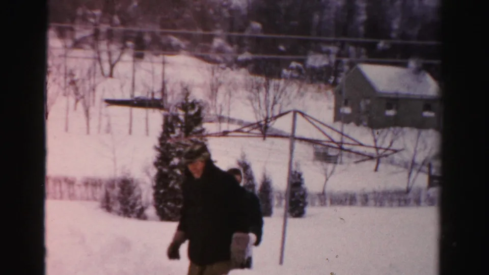 An adult in a dark coat faces toward the camera in a snowy Albany backyard, with a clothesline, shrubs, and outbuilding behind.