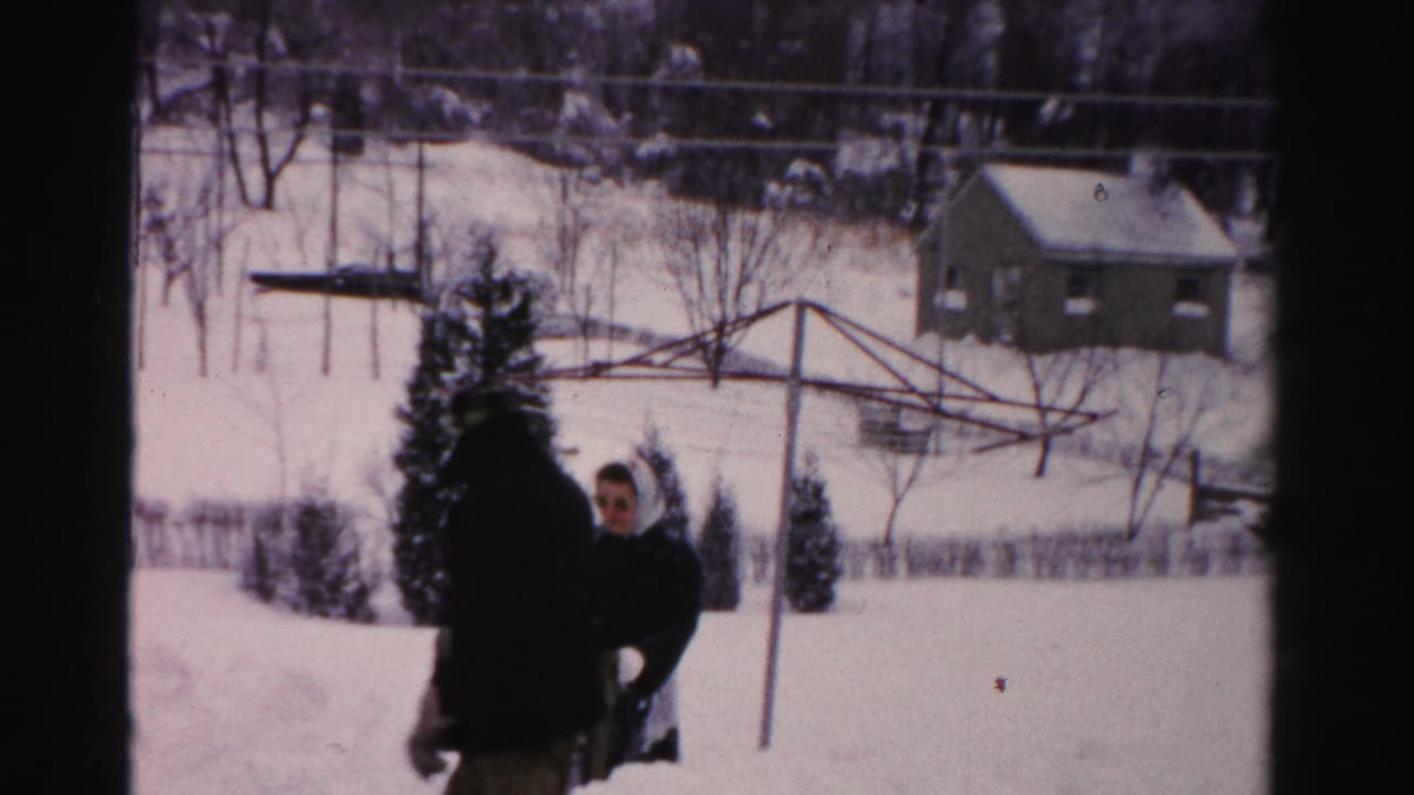 1950s Albany home-movie frame showing two adults in winter clothing in a snow-covered yard with bare trees and a small house beyond.