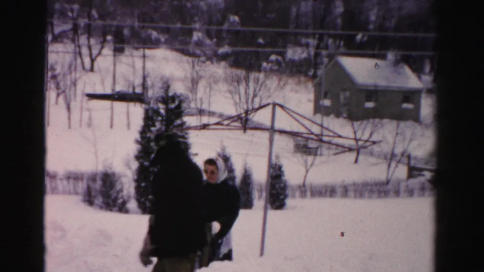 1950s Albany home-movie frame showing two adults in winter clothing in a snow-covered yard with bare trees and a small house beyond.