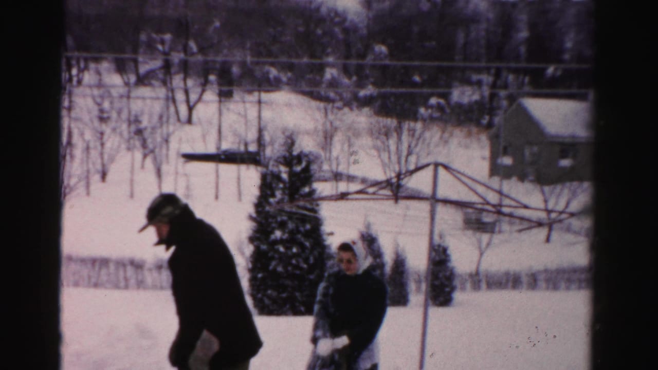 A man in a dark coat walks through snow while a woman stands nearby in the yard.