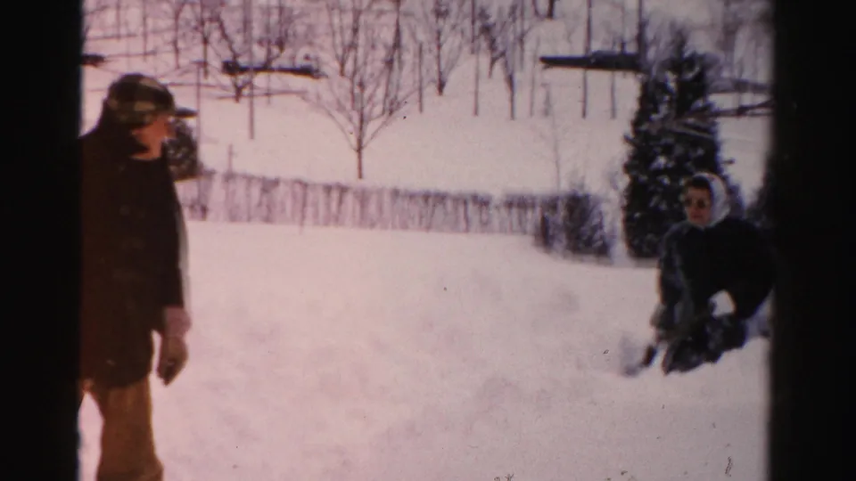 Home-movie frame with a man partly at left and a bundled woman low in the snow, with a snowy yard and trees behind them.