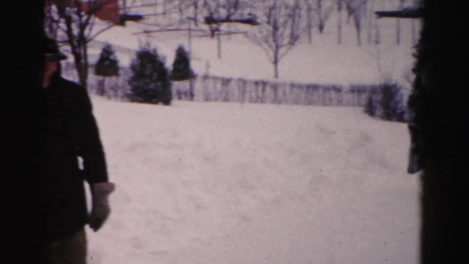 A dark-coated adult passes close to the camera in a snowy Albany yard, partially obscuring the winter landscape behind.