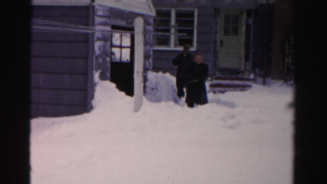 Two men stand in deep snow outside a house with siding, windows, and a front door.
