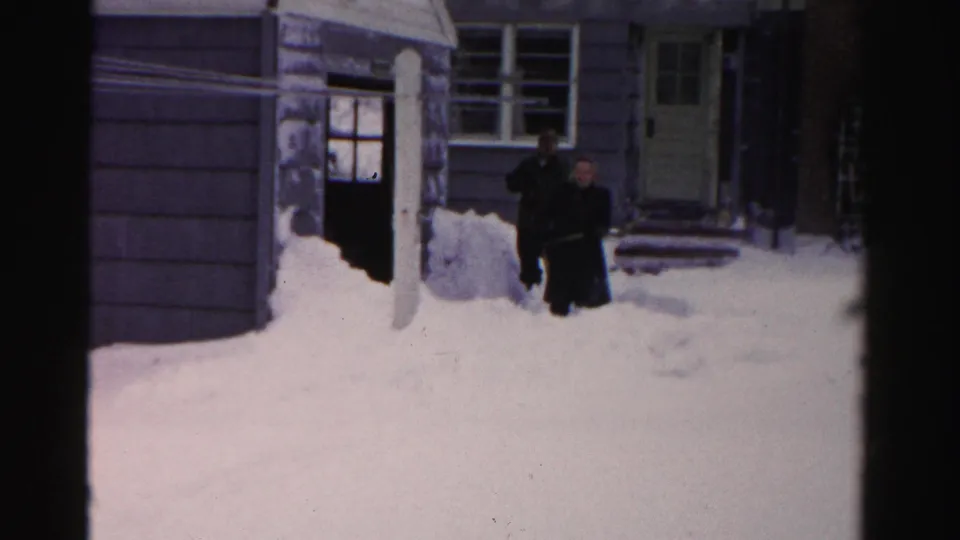 Two men in dark winter coats stand in deep snow outside an Albany residence in archival 8mm footage dated 1956.