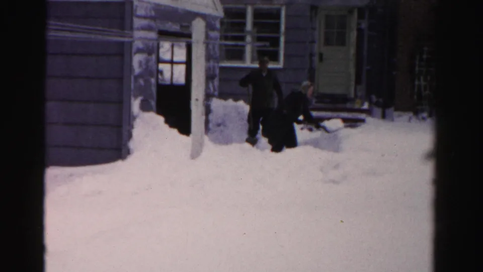 Two men in dark coats work through heavy snow near the entrance of an Albany home in 1956 8mm footage.
