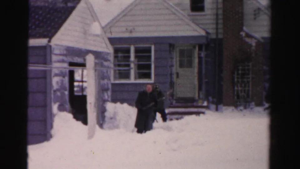 A man in a dark coat works in deep snow near the front steps of an Albany residence in archival 8mm footage.