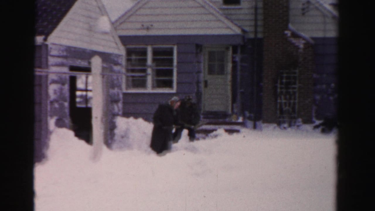 In Albany, New York home-movie footage, two men in dark winter coats appear to clear snow outside a light-sided house.