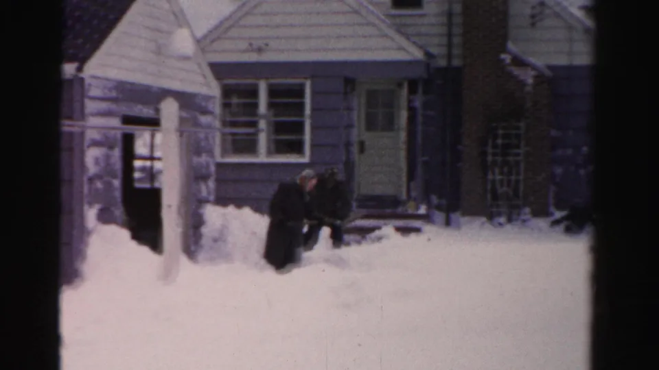 In Albany, New York home-movie footage, two men in dark winter coats appear to clear snow outside a light-sided house.