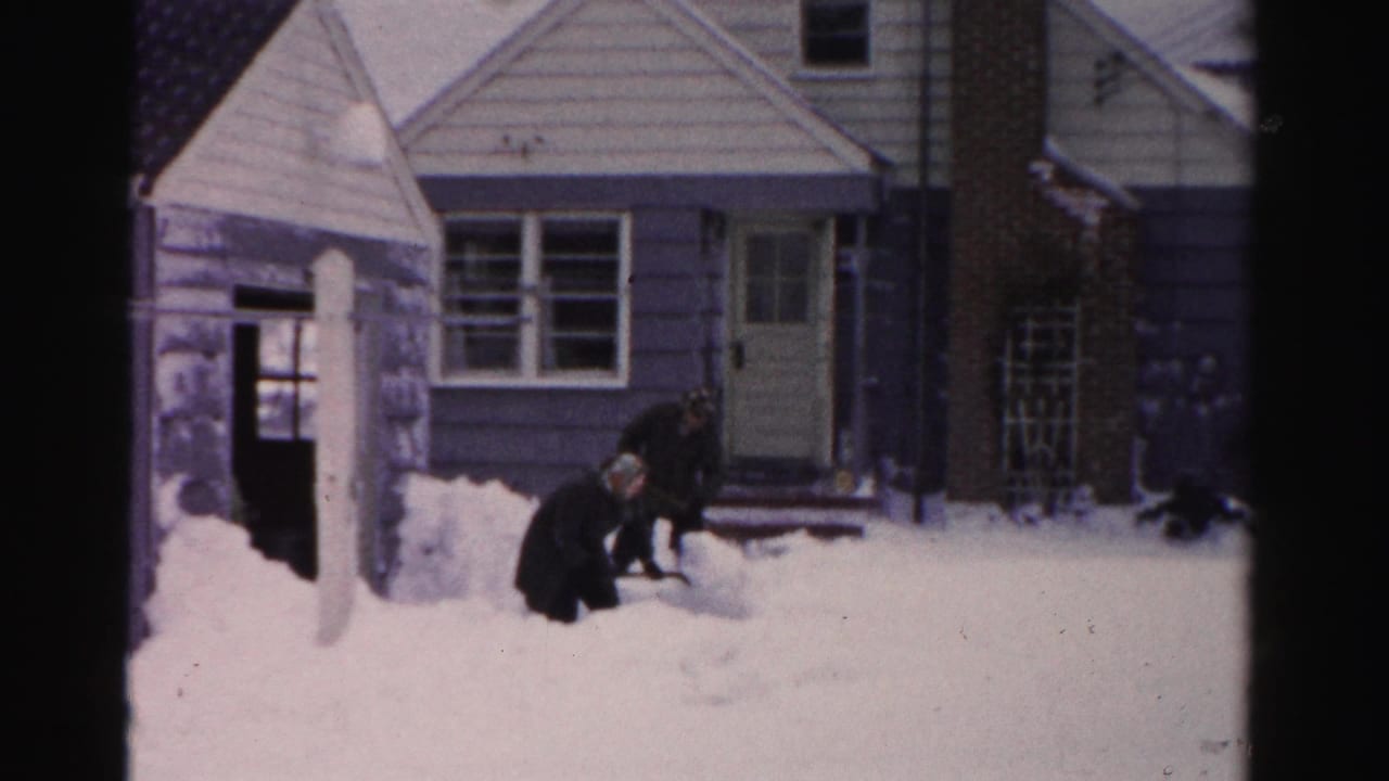 Two men bend over a snowbank outside a house with front steps and windows.