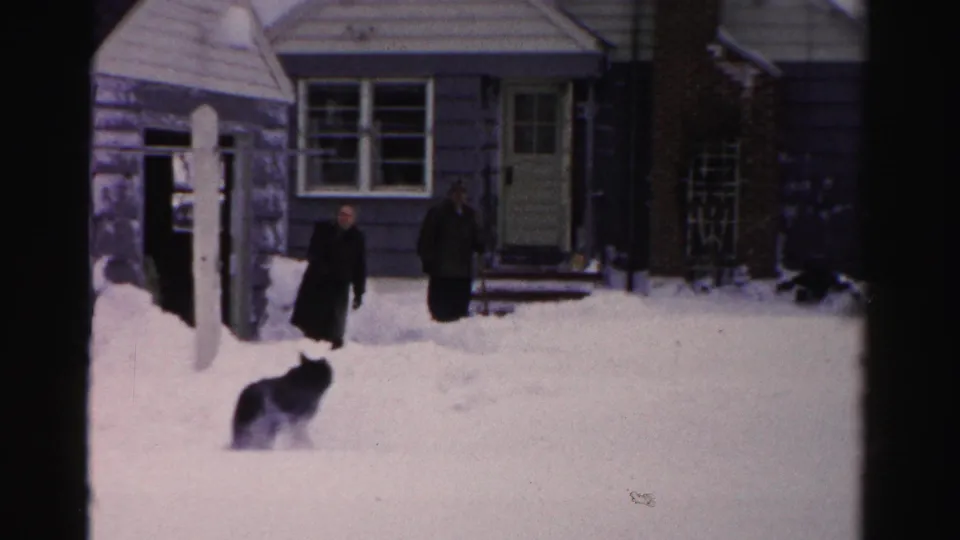 Two bundled men stand outside the Albany house in snow while a dog enters the foreground of the home-movie frame.