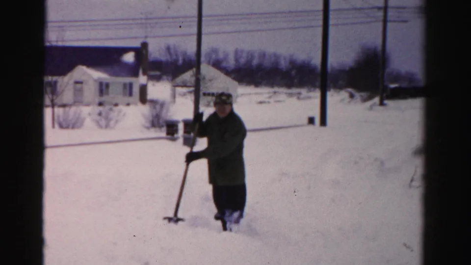 Adult in winter coat shovels heavy snow in a residential front yard; wooden utility poles, bare deciduous trees, and mid-century suburban homes visible in background, c. mid-1950s.
