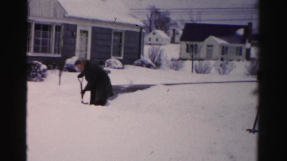 Adult in winter coat bent low shoveling deep snow; mid-century suburban homes, bare trees, and a garage-style outbuilding visible in background, c. mid-1950s Albany, NY.