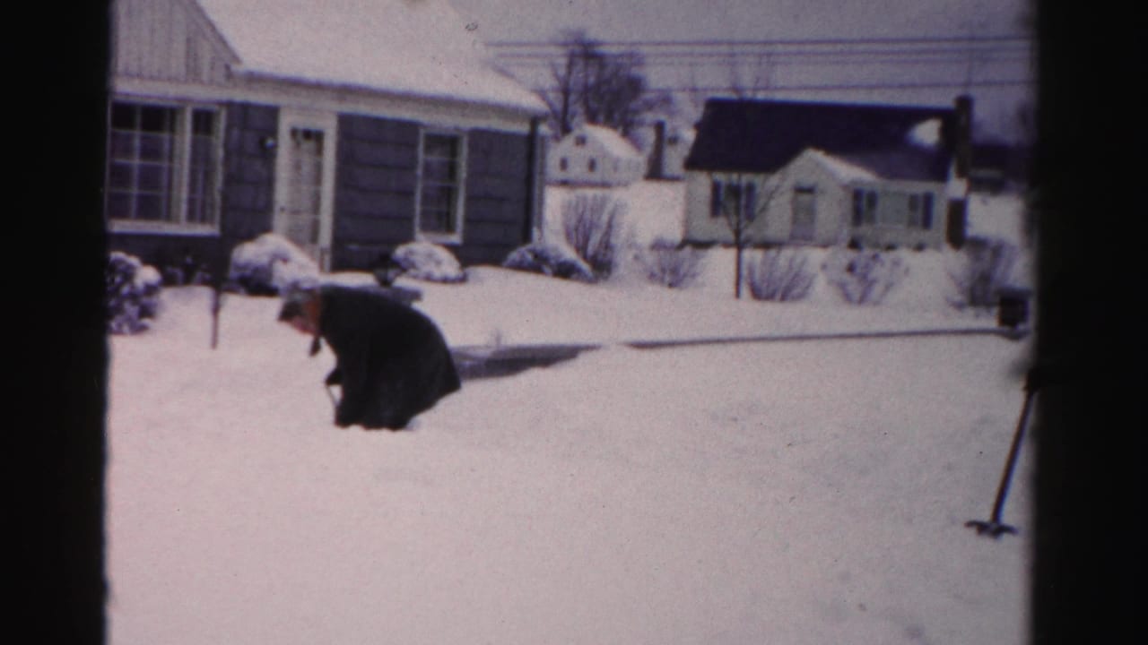 Man shoveling heavy snow in front of mid-century ranch-style homes, Albany, New York area, circa mid-1950s, captured on 8mm home movie film.