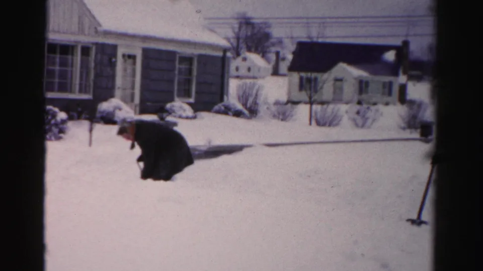 Man shoveling heavy snow in front of mid-century ranch-style homes, Albany, New York area, circa mid-1950s, captured on 8mm home movie film.