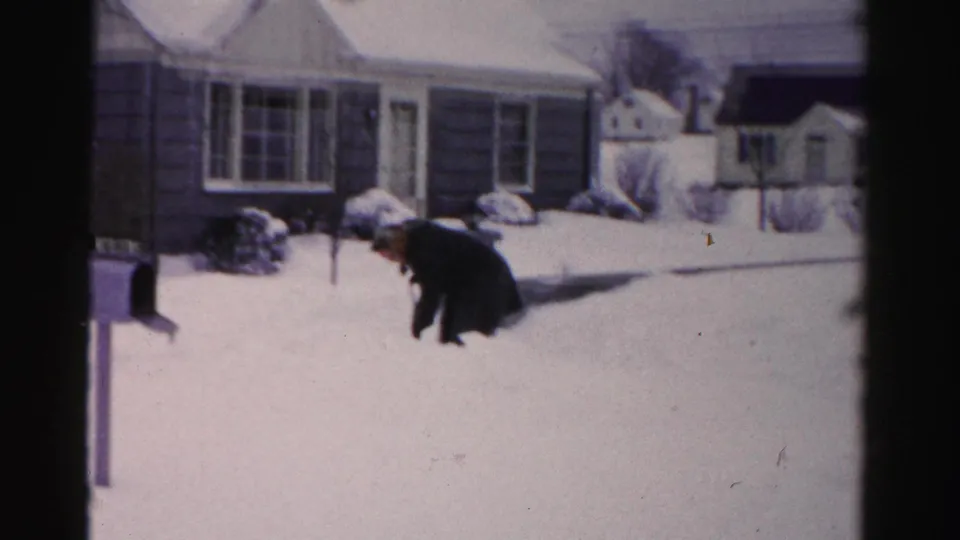 Man shoveling snow in suburban front yard; mid-century dark-sided ranch home with white trim and mailbox post; Albany, New York area, circa mid-1950s.