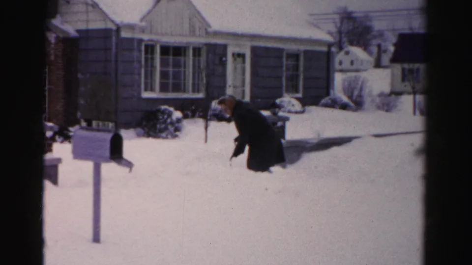 Man clearing deep snow from front yard; mid-century ranch home with dark horizontal siding; mailbox post prominent in foreground; Albany, New York area, mid-1950s.