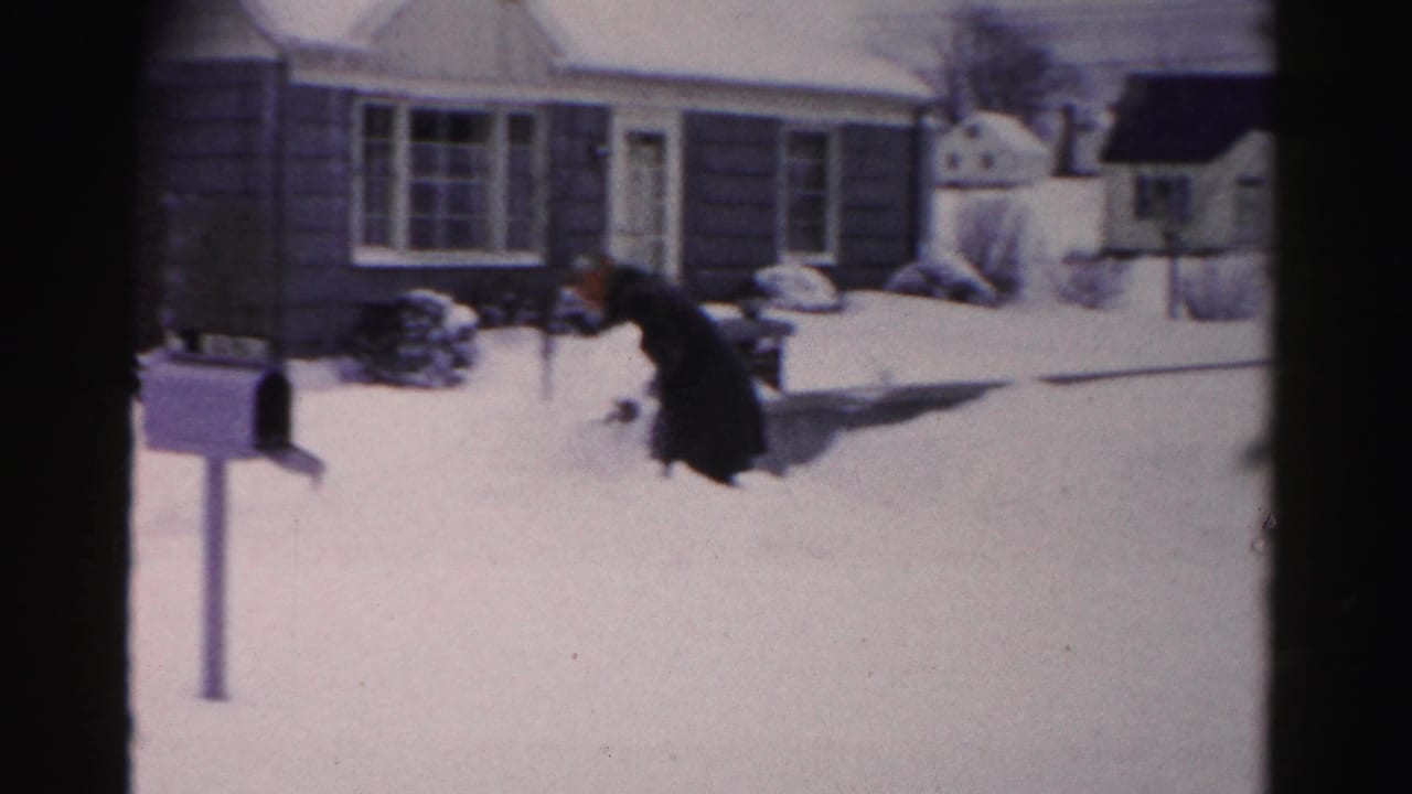 Man in heavy dark coat crouching to shovel deep snow in a mid-1950s suburban yard; single-story house and mailbox post visible behind him.