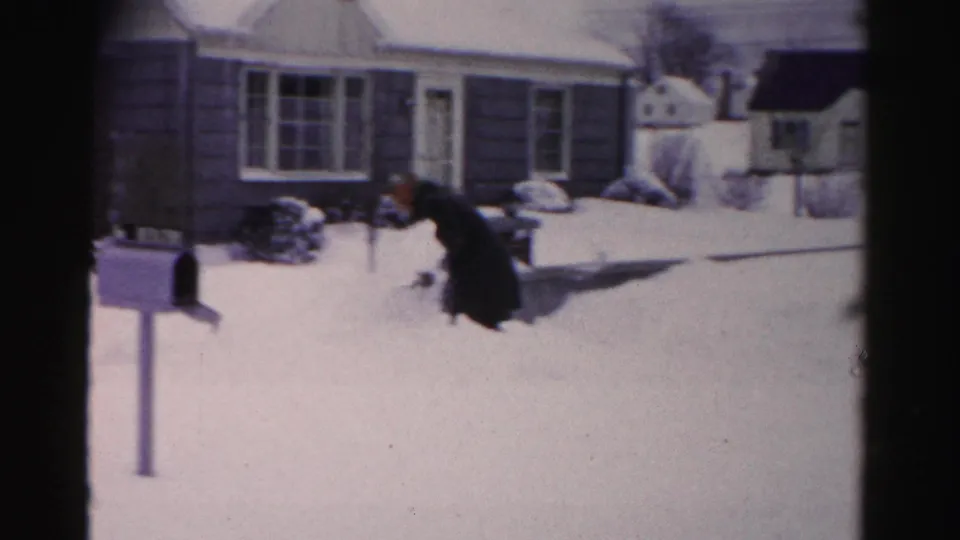 Man in heavy dark coat crouching to shovel deep snow in a mid-1950s suburban yard; single-story house and mailbox post visible behind him.
