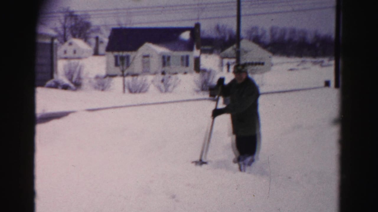 Man in dark coat and hat standing with snow shovel in a mid-1950s suburban neighborhood; multiple houses, bare trees, and utility poles visible in background.