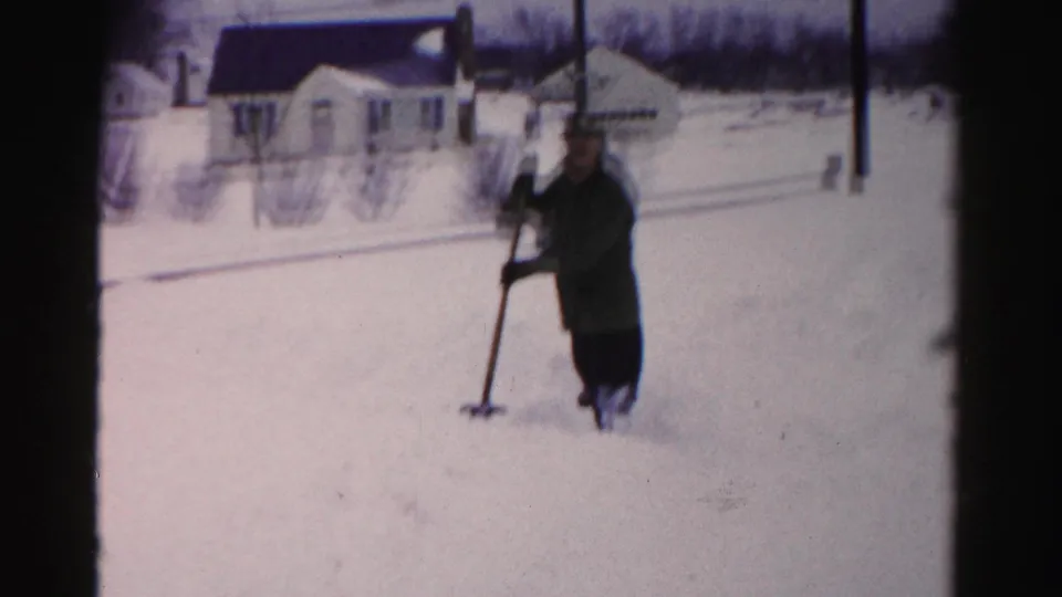 Man in dark winter coat holding a snow shovel, positioned closer to camera; mid-1950s white suburban house with dark roof and utility poles behind him.