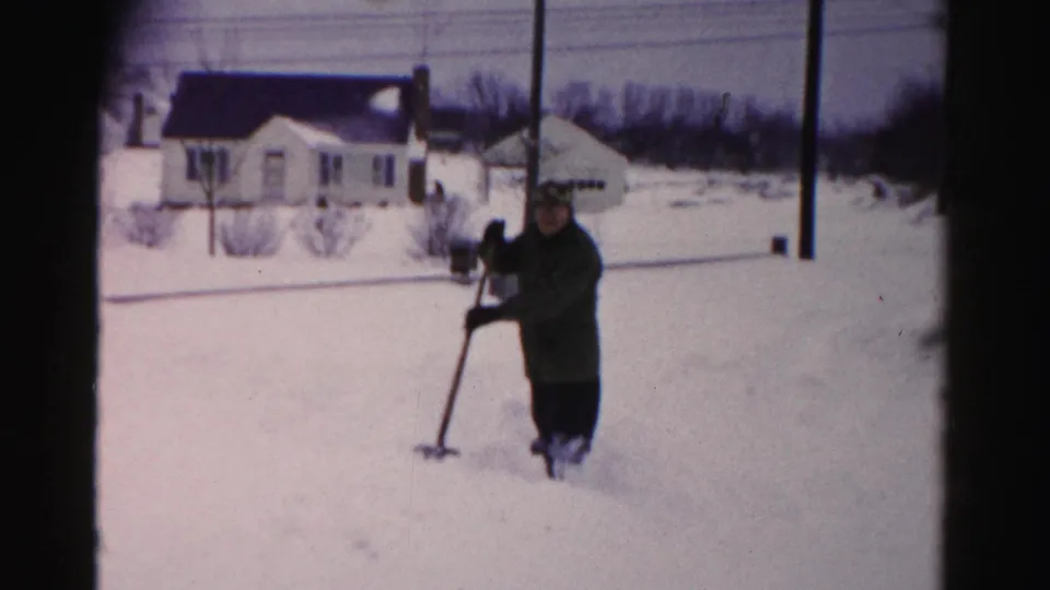 Man in dark coat and hat holding snow shovel in a snow-covered 1950s suburban yard; neighboring house and telephone poles with wires visible behind him.