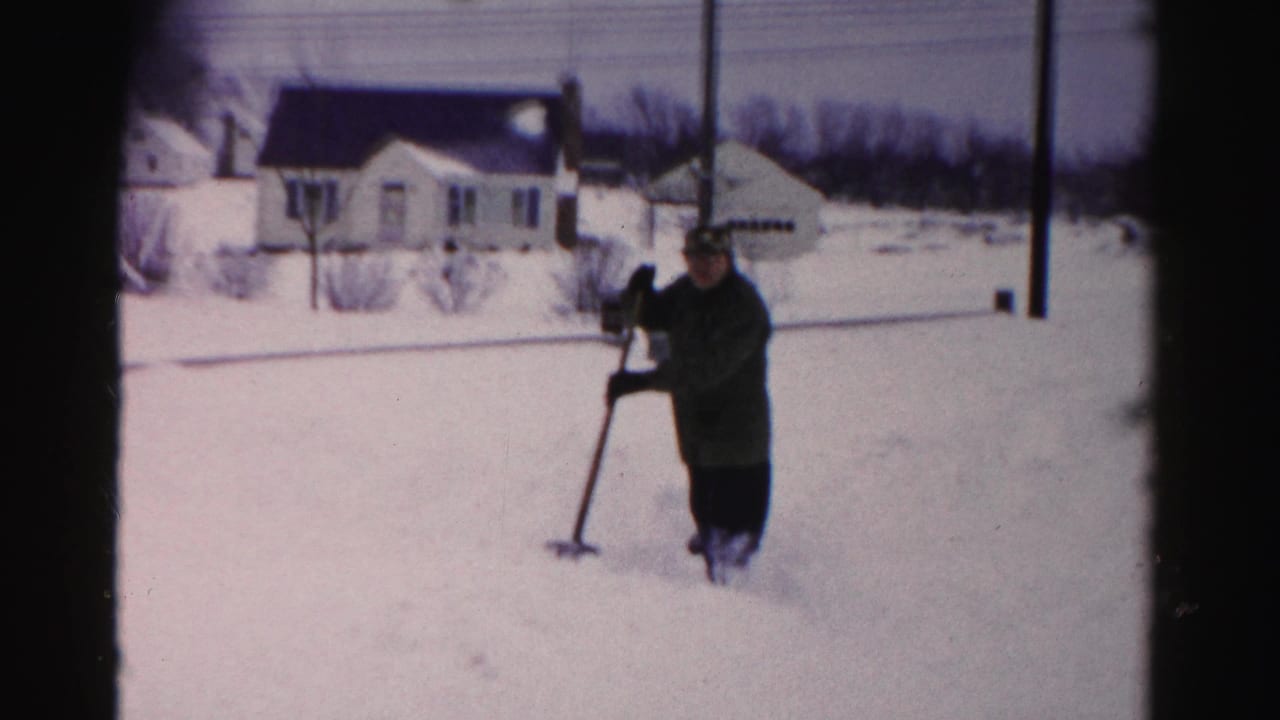 Man in dark coat shoveling deep snow in a suburban yard, white house with dark roof in background, mid-century 8mm film.