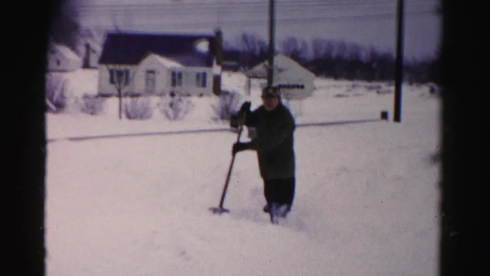 Man in dark winter coat and hat shoveling heavy snow in a suburban yard, Albany, New York area, circa mid-1950s, captured on 8mm home movie film.