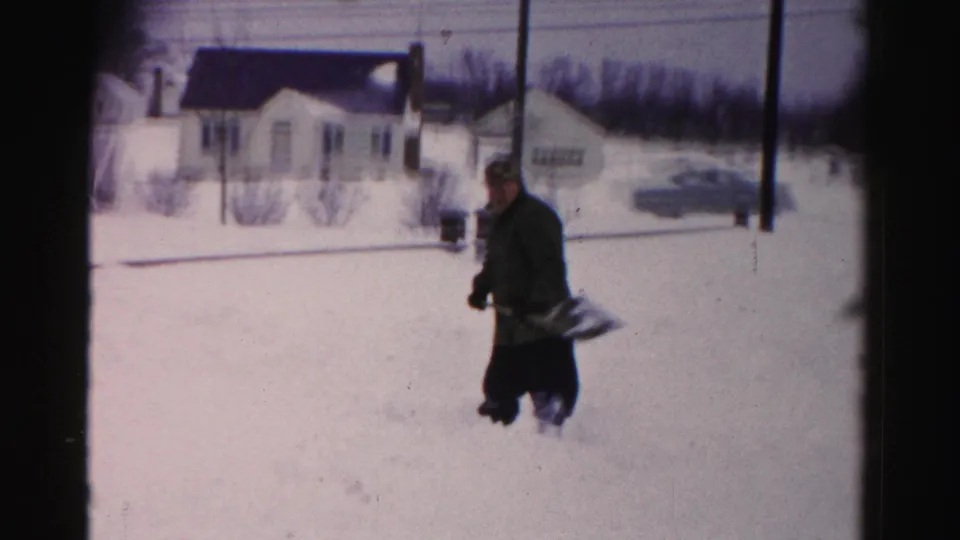 Medium shot of a man in dark winter coat shoveling snow, white postwar ranch-style house in background, 8mm home movie, Albany, New York area, c. mid-1950s.