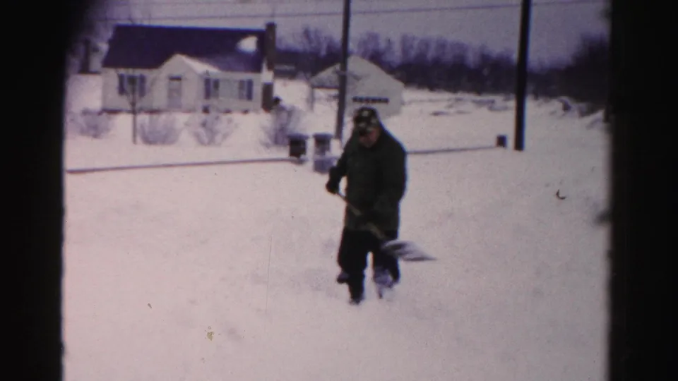 Man in dark winter coat throwing snow from shovel in suburban yard, Albany, New York area, circa mid-1950s, 8mm home movie footage.