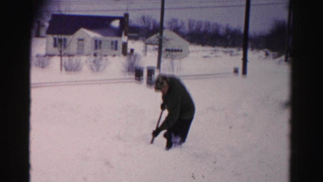 Man in dark coat bends forward shoveling snow in a residential yard, white house and utility poles visible behind him.
