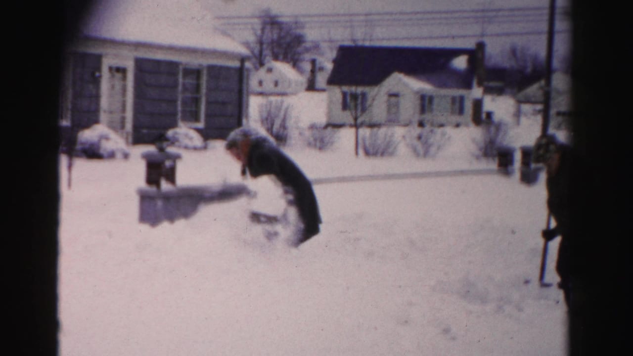 The same man bends close to snow level near a metal mailbox in what may be a driveway area of a mid-1950s suburban home on 8mm film.
