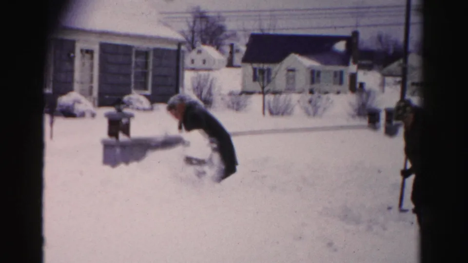 The same man bends close to snow level near a metal mailbox in what may be a driveway area of a mid-1950s suburban home on 8mm film.