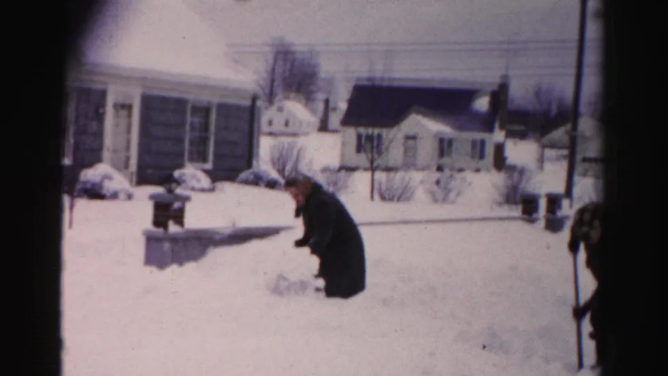A man crouches low in heavy snow in a mid-1950s suburban neighborhood; a second partially visible figure enters from the right edge of the 8mm frame.