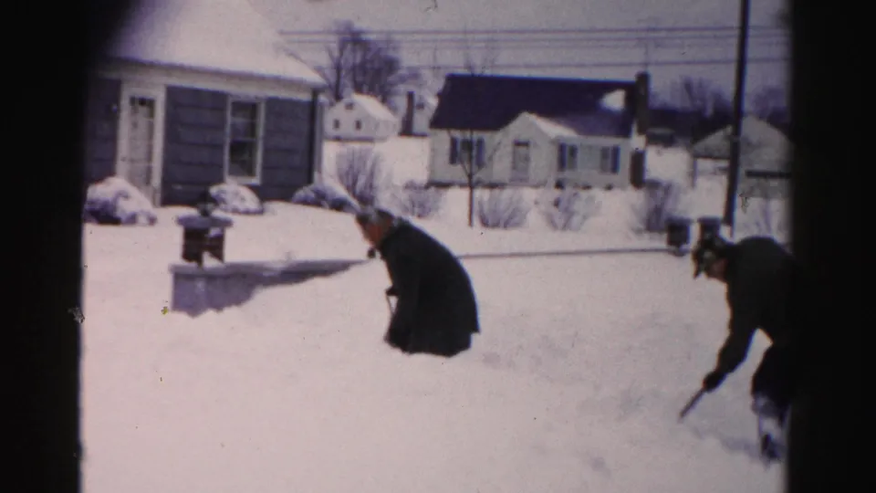 Two people in winter coats appear in a snow-covered suburban yard in what may be Albany, New York, circa mid-1950s, captured on 8mm home-movie film.