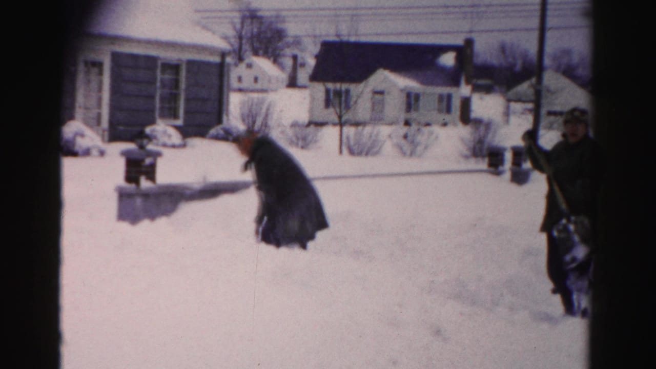 Two residents in dark coats shoveling snow in a mid-1950s suburban neighborhood, possibly Albany, New York, with cape cod and ranch-style houses behind them.