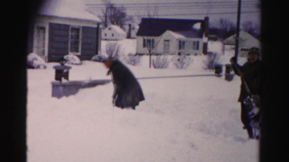 Two residents in dark coats shoveling snow in a mid-1950s suburban neighborhood, possibly Albany, New York, with cape cod and ranch-style houses behind them.