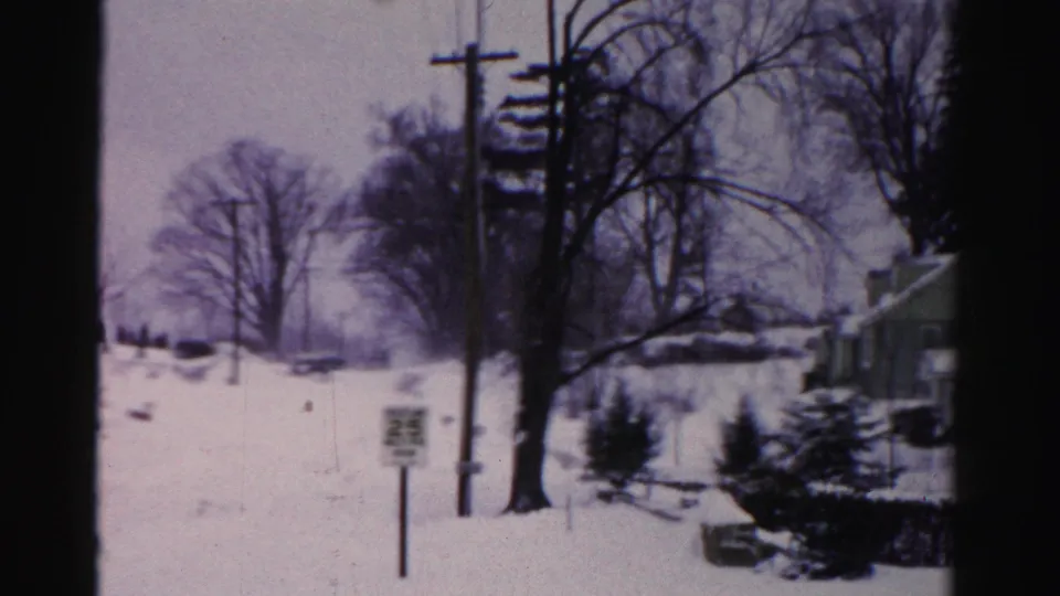 Pan across a snow-covered mid-1950s suburban street, 25 mph sign prominent in foreground, bare deciduous trees and utility poles overhead.