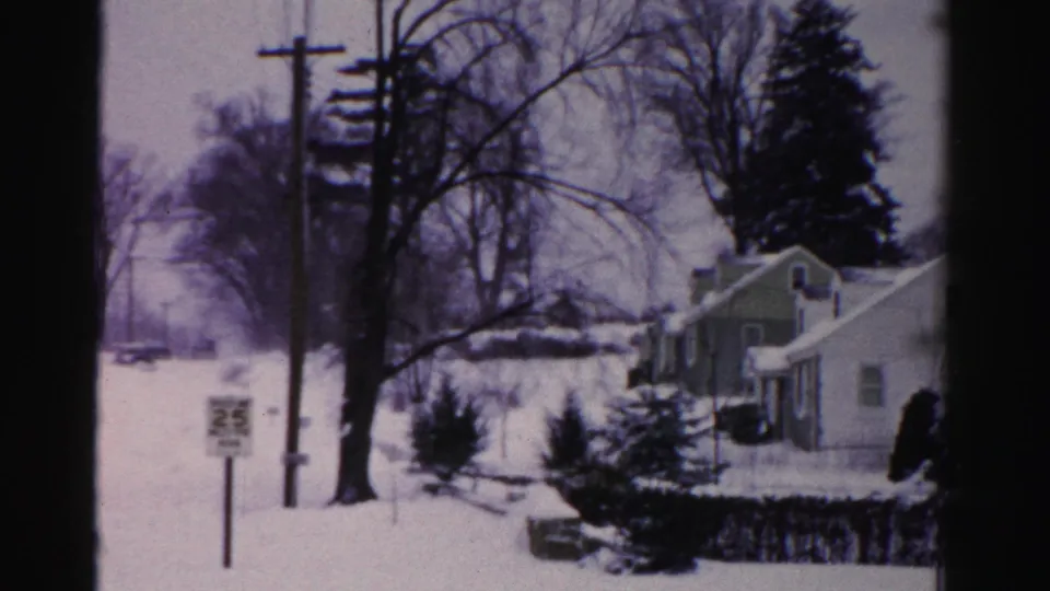 End-of-clip view of a snow-covered suburban street, possibly Albany, New York mid-1950s, with a light-colored house, evergreen trees, and a 25 mph sign visible.