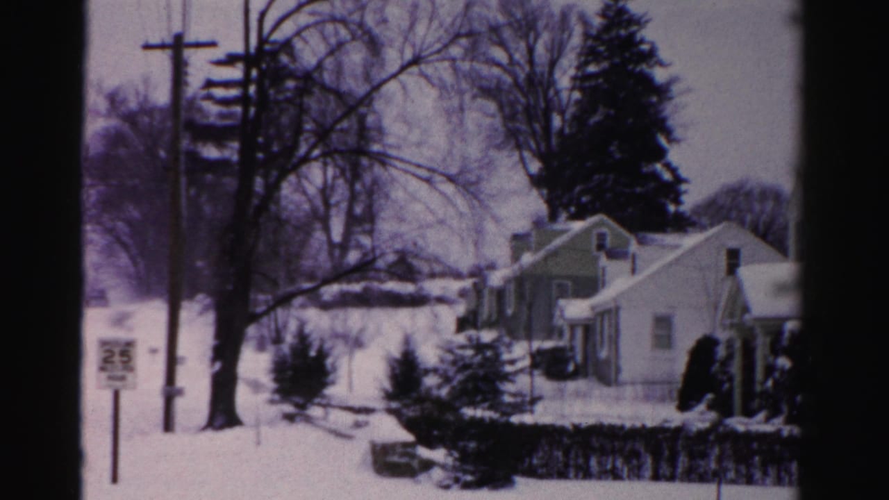 8mm home movie frame circa mid-1950s: snow-blanketed Albany, New York suburban street with modest one-story homes, bare trees, and a roadside sign on a utility pole.